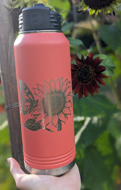 32 oz coral Polar Camel water bottle held outdoors with laser engraved sunflower and butterfly design, real sunflowers in background